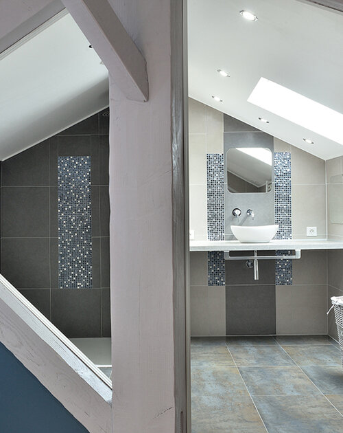 Suite parentale sous les combles avec mise en valeur des poutres. Création de rangements sous les rampants. Magnifique salle de douche épurée et raffinée. L'ensemble de l'espace est ouvert pour une très belle perspective. Les tons de dégradés de gris, bleu, chrome, sont réchauffés par le bois et les briques rouges.